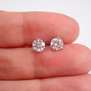May include: A pair of round, clear diamond stud earrings. Each earring features a sparkling, faceted diamond held in place by a silver-colored setting. The earrings are displayed against a light background.