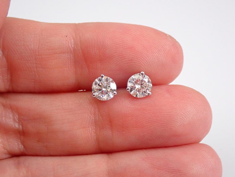 May include: A pair of round, clear diamond stud earrings. Each earring features a sparkling, faceted diamond held in place by a silver-colored setting. The earrings are displayed against a light background.