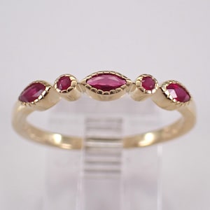 May include: A gold ring featuring five vibrant red gemstones. The gemstones are set in a delicate, alternating pattern, with a marquise-cut stone at the center and round and oval stones on either side. The ring has a polished, smooth band.