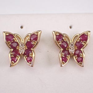 Puede incluir: Un par de pendientes de mariposa dorados. Cada pendiente presenta un diseño de mariposa con piedras preciosas de rubí y un pequeño acento de diamante en el centro. Los pendientes están sobre un fondo blanco.