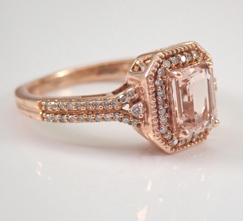 May include: A rose gold engagement ring with a large, light pink, emerald-cut gemstone surrounded by a halo of smaller diamonds. The band is also set with smaller diamonds.