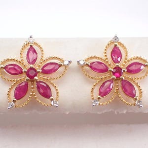 May include: A pair of flower-shaped stud earrings. Each earring features a gold-colored frame with a beaded edge, holding five oval-cut, deep red gemstones and a central round gemstone. Small white gemstones are set at the tips of the petals.