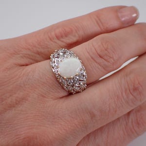 May include: An oval opal ring with an elaborate silver setting. The ring has a large, milky white opal stone at its centre, encircled by smaller, clear stones. The setting features an intricate filigree design.