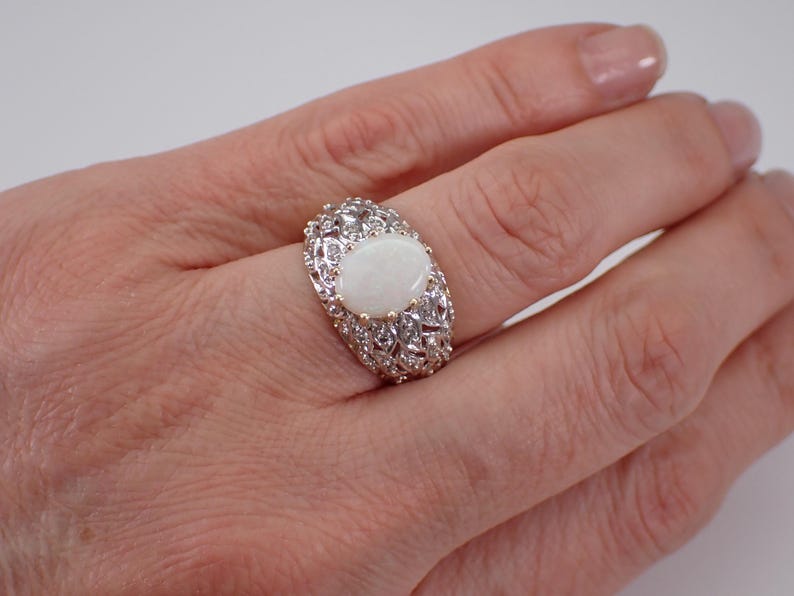 May include: An oval opal ring with an elaborate silver setting. The ring has a large, milky white opal stone at its centre, encircled by smaller, clear stones. The setting features an intricate filigree design.