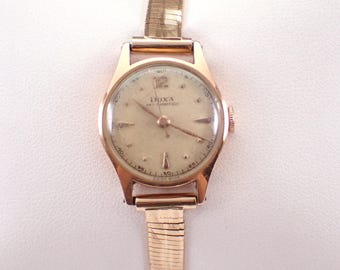 Könnte beinhalten: Eine vintage goldfarbene Armbanduhr mit cremefarbenem Zifferblatt. Die Uhr trägt den Markennamen "Doxa" und "Anti-Magnetique" auf dem Zifferblatt. Die Uhr hat ein goldfarbenes Armband und eine kleine Krone. Das Zifferblatt hat schwarze Ziffern und Markierungen.