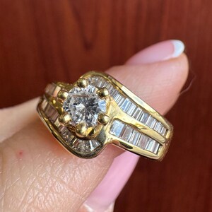May include: A gold ring featuring a central round brilliant-cut stone held by six prongs, flanked by two rows of channel-set baguette-cut stones that follow a bypass design along the band, creating a layered appearance with clear gemstones.