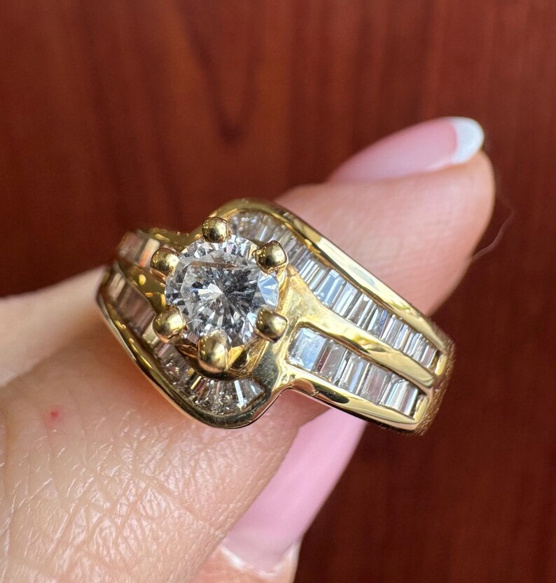 May include: A gold ring featuring a central round brilliant-cut stone held by six prongs, flanked by two rows of channel-set baguette-cut stones that follow a bypass design along the band, creating a layered appearance with clear gemstones.