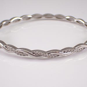 May include: A silver-toned bangle bracelet with a twisted design. The bracelet features small, sparkling stones set along the twisted sections. The clasp is visible on one side, and the bracelet is displayed on a white surface.