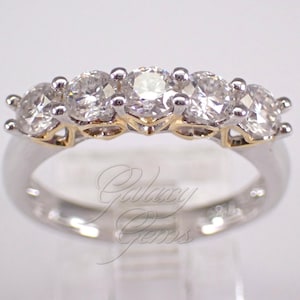 Può includere: Un anello a cinque pietre con gemme chiare a taglio brillante rotondo, incastonate in una fascia di metallo color argento con accenti color oro sotto le pietre. Il testo Galaxy Gems è visibile in carattere corsivo sulla parte inferiore dell'anello.