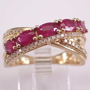 May include: A gold crossover ring featuring four oval-cut deep red rubies set in a row, accented by two intersecting bands encrusted with small clear white diamonds. The metal band has a polished finish and a structured, multi-layered design.