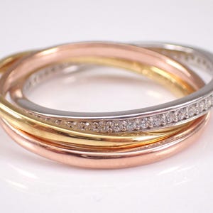 May include: A three-band ring with interwoven bands in gold, rose gold, and silver. The silver band is set with small gemstones. The ring design represents unity and elegance.