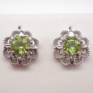 May include: A pair of silver earrings with a green gemstone in the center of each earring. The earrings have a delicate floral design.