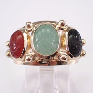 May include: A gold ring featuring three oval-shaped gemstones: a red carnelian, a light green stone, and a black onyx. The ring has a detailed design with small gold accents around the gemstones, showcasing an antique or vintage style.