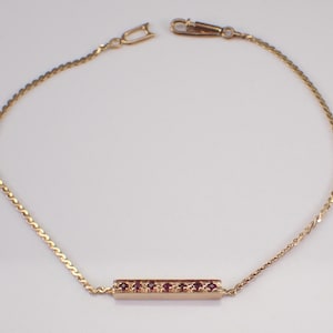 May include: A gold-colored bracelet with a bar-shaped centerpiece adorned with small, red gemstones. The bracelet features a delicate chain design and a clasp for secure fastening. The jewelry is displayed against a plain white background.