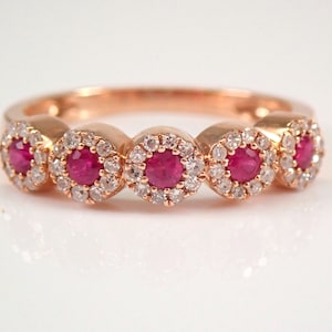 May include: Rose gold ring featuring five round, vibrant red gemstones, each encircled by a halo of smaller, sparkling white stones. The band is smooth and polished, creating a classic and elegant design.