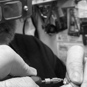 May include: A close-up of a jeweler using a tool to work on a piece of jewelry. The jeweler is wearing a magnifying glass and is focused on their work.