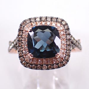 Könnte beinhalten: Ein Ring aus Roségold mit einem dunkelblauen Edelstein im Kissenschliff. Der Mittelstein ist von einem doppelten Halo aus kleinen, runden braunen Diamanten umgeben. Die Ringschiene hat ein geteiltes Design mit braunen Diamanten.