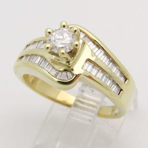 May include: A yellow gold ring featuring a central round brilliant-cut diamond held in a six-prong setting. The band is accented with two rows of channel-set baguette-cut diamonds that follow the curved, bypass-style design of the metal.
