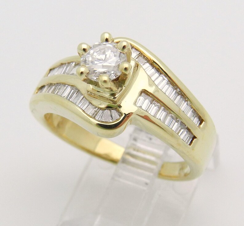 May include: A yellow gold ring featuring a central round brilliant-cut diamond held in a six-prong setting. The band is accented with two rows of channel-set baguette-cut diamonds that follow the curved, bypass-style design of the metal.