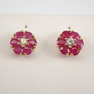 Pode incluir: Um par de brincos de ouro com um design de flor. Cada brinco apresenta um grupo de pedras de rubi vermelho que envolvem um único diamante branco.