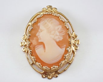 Könnte beinhalten: Ovale Cameo-Brosche mit einem pfirsichfarbenen Relief eines Frauenprofils. Der Cameo ist in einem goldfarbenen Filigranrahmen mit dekorativen Verzierungen gefasst. Ein klassisches Schmuckstück.