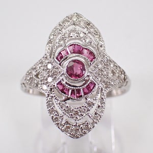 May include: A silver-toned ring with a central cluster of pink gemstones surrounded by smaller clear stones. The ring has an ornate, Art Deco-inspired design with a symmetrical shape and intricate detailing.