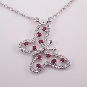 May include: A silver-toned butterfly pendant necklace featuring a delicate openwork frame encrusted with small clear stones. The butterfly wings are accented with nine round, deep red gemstones set within the metal structure on a fine chain.