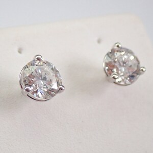 May include: A pair of sparkling diamond stud earrings. Each earring features a large, round, brilliant-cut diamond set in a silver-colored, three-prong setting. The diamonds are clear and reflect light, creating a brilliant sparkle.