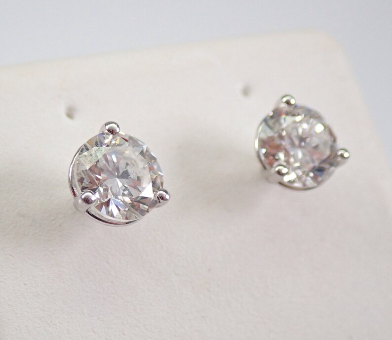 May include: A pair of sparkling diamond stud earrings. Each earring features a large, round, brilliant-cut diamond set in a silver-colored, three-prong setting. The diamonds are clear and reflect light, creating a brilliant sparkle.