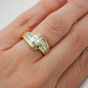 May include: A yellow gold ring featuring a central round brilliant-cut diamond held in a four-prong setting, flanked by two rows of channel-set baguette-cut diamonds that follow the curved, bypass-style design of the polished gold band.
