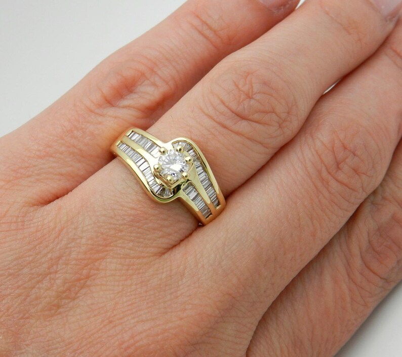 May include: A yellow gold ring featuring a central round brilliant-cut diamond held in a four-prong setting, flanked by two rows of channel-set baguette-cut diamonds that follow the curved, bypass-style design of the polished gold band.