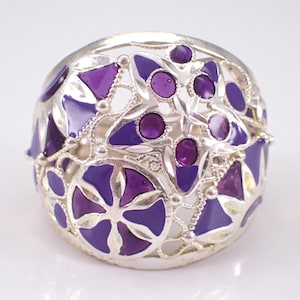 Könnte beinhalten: Ein breiter Silberring mit einem komplizierten durchbrochenen Design, das mit leuchtend violetten Emaille-Akzenten gefüllt ist. Die Metallarbeit zeigt eine Vielzahl geometrischer und floraler Formen, die eine detaillierte, strukturierte Oberfläche bilden.