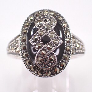 Könnte beinhalten: Ovale Silberring mit einem schwarzen Onyx-Mittelstein. Der Ring ist mit Markasitsteinen um den Onyx und auf dem Band besetzt. Die Markasitsteine sind klein und rund und verleihen dem Ring Glanz.