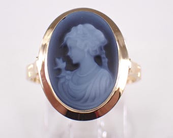 May include: A gold ring featuring an oval cameo with a blue silhouette of a woman. The ring has a detailed gold band and a classic design. The cameo depicts a woman holding a flower.