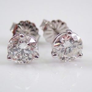 May include: A pair of diamond stud earrings. Each earring features a large, round, brilliant-cut diamond set in a three-prong setting. The earrings are displayed on a white surface, reflecting light and showcasing their sparkle.