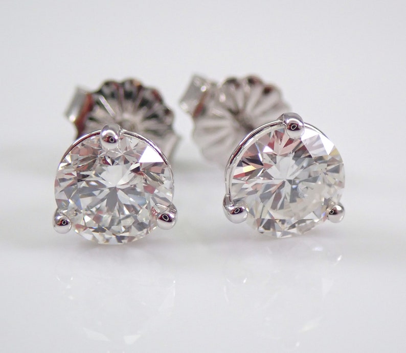 May include: A pair of diamond stud earrings. Each earring features a large, round, brilliant-cut diamond set in a three-prong setting. The earrings are displayed on a white surface, reflecting light and showcasing their sparkle.