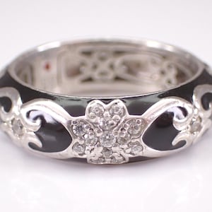 Könnte beinhalten: Ein silberfarbener Bandring mit einer Einlage aus poliertem schwarzem Emaille. Der Ring ist mit sich wiederholenden silbernen Schnörkelmustern und einem zentralen Blumenmotiv verziert, das mit kleinen, klaren, runden Steinen besetzt ist.