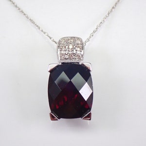May include: A silver chain necklace with a pendant featuring a large, dark red gemstone surrounded by a square of smaller, clear gemstones.