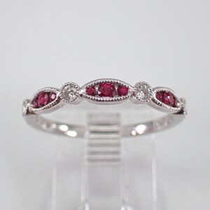 May include: A delicate silver ring featuring alternating oval-shaped settings with vibrant red gemstones and round, clear gemstones. The ring's design is symmetrical, with a repeating pattern of red and clear stones.