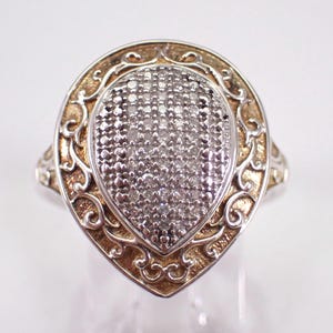 May include: A silver ring with a teardrop-shaped face. The center is filled with small, sparkling stones. The ring has a gold-colored filigree border and a silver band. The ring is set against a white background.