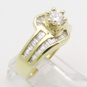 May include: A yellow gold ring featuring a central round brilliant-cut diamond held in a six-prong setting. The band is accented with multiple channel-set baguette-cut diamonds arranged in two parallel rows along the shank of the ring.