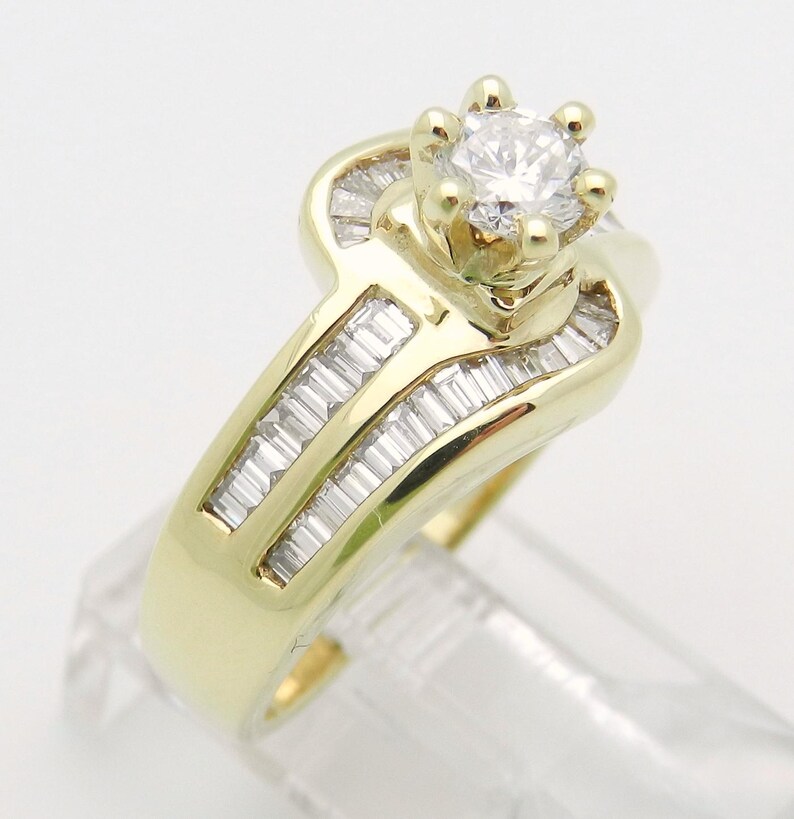 May include: A yellow gold ring featuring a central round brilliant-cut diamond held in a six-prong setting. The band is accented with multiple channel-set baguette-cut diamonds arranged in two parallel rows along the shank of the ring.
