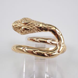 Puede incluir: Un anillo de metal dorado con forma de serpiente enroscada. El anillo presenta una cabeza detallada y texturizada con ojos y escamas definidos, y un cuerpo liso y curvado que se envuelve para formar un diseño de banda abierta y ajustable.