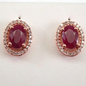 Op de afbeelding: Een paar roségouden oorstekers. Elke oorbel heeft een grote, ovaalvormige, dieprode robijn, omringd door een halo van kleine, heldere, rond geslepen diamanten. De oorbellen zijn geplaatst tegen een witte achtergrond.