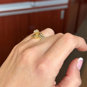 May include: A gold ring featuring a central round-cut diamond held in a prong setting, flanked by a row of channel-set baguette diamonds along the band. The metal band has a polished finish and a curved design that tapers toward the center stone.