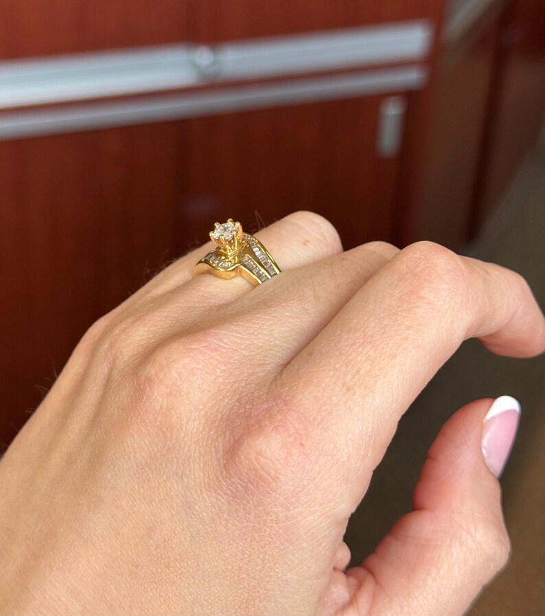May include: A gold ring featuring a central round-cut diamond held in a prong setting, flanked by a row of channel-set baguette diamonds along the band. The metal band has a polished finish and a curved design that tapers toward the center stone.