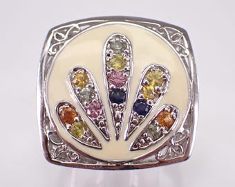 May include: A silver-toned ring featuring a square frame with intricate filigree corners. The center is a circular cream-colored enamel inlay decorated with five radiating fan-shaped prongs, each set with small, multi-colored round gemstones.