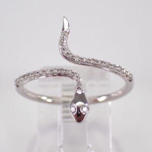 May include: A silver snake ring with a curved design. The ring features a snake head and tail, both set with small, sparkling stones. The snake's body curves around the finger, creating a unique and elegant piece of jewellery.