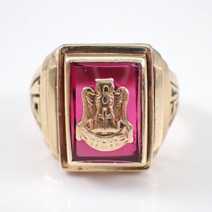 May include: A gold ring with a red stone featuring a crest with a bird and the word "ALPHA" inscribed.