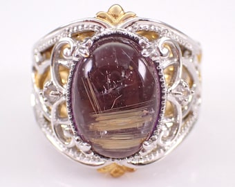 May include: A silver and gold ring featuring an oval, dark purple gemstone with golden inclusions. The ring has an ornate filigree design around the gemstone and gold accents.